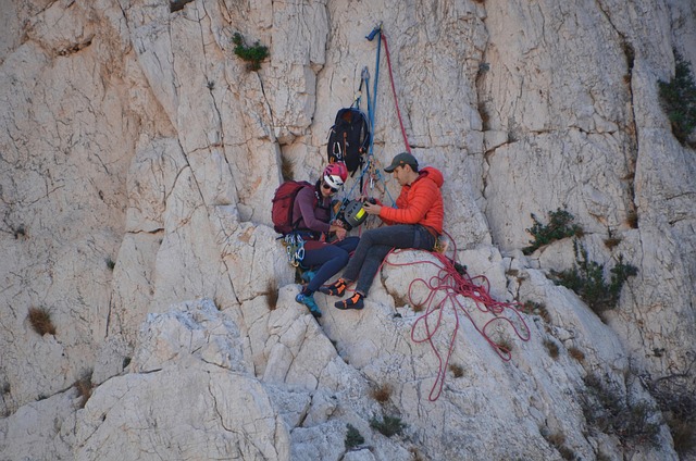 Two climbers resting on a rocky cliff face, wearing helmets and harnesses with ropes and gear nearby.