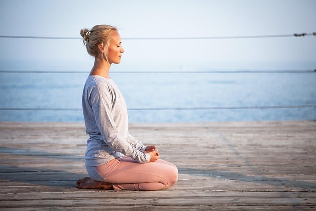 Woman meditating in a cross-legged pose on a wooden pier, eyes closed, overlooking a calm sea at dusk.